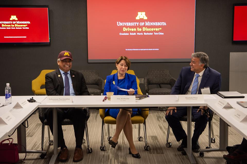Three people sitting at table, one wearing a UMN hat and smiling in front of screens saying UMN.